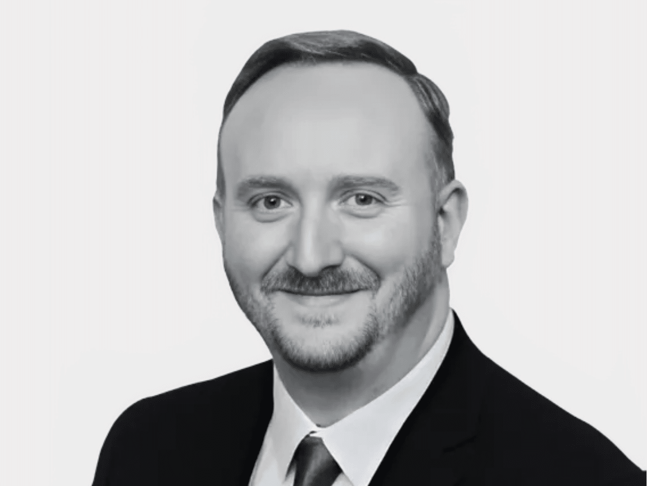 Black and white headshot of Kevin Deegan, Policy & Communications Lead, wearing a dark suit, white shirt, and tie, smiling.